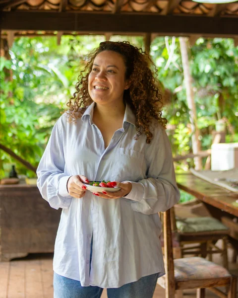 Mulher sorridente de camisa azul segurando uma tigela de frutas numa varanda com plantas