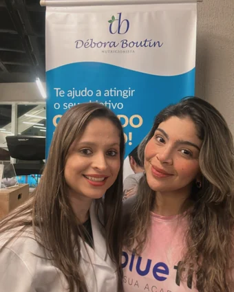 Débora de jaleco sorrindo ao lado de uma mulher de camiseta rosa da Bluefit, diante do banner dela
