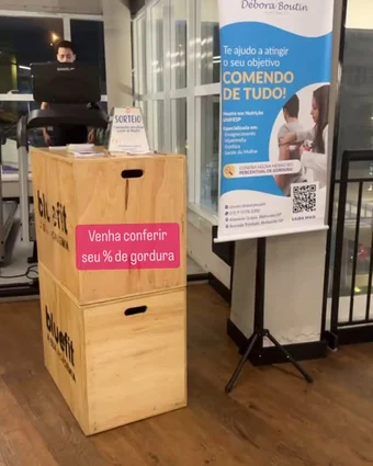 Estande com o banner da Débora Boutin e caixotes de madeira numa academia